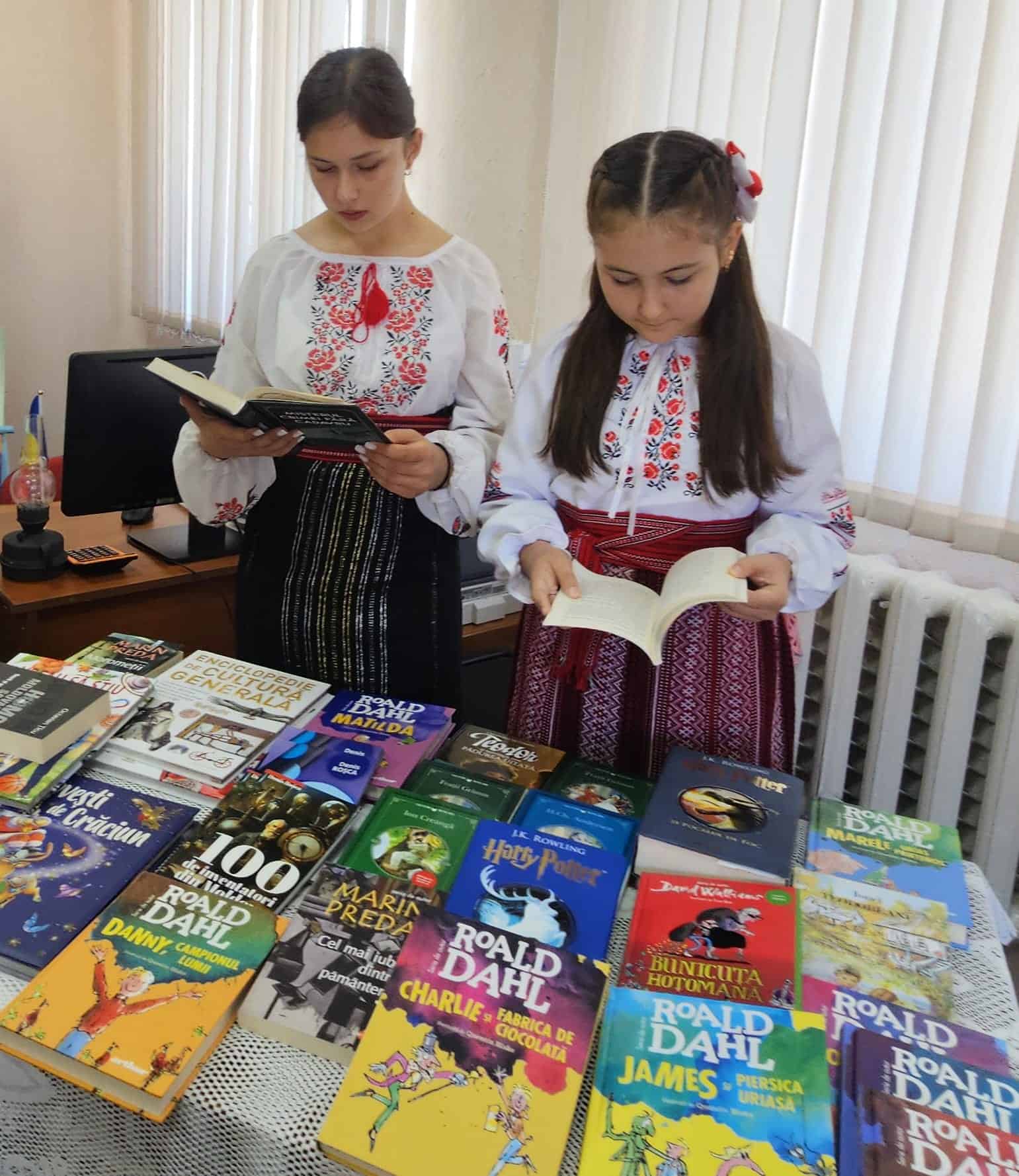 Twee Moldavische meisjes in traditionele kleding lezen Roald Dahl-boeken tijdens SMHO-hulpactie in schoolbibliotheek, ter bevordering van leesplezier en educatie in Oost-Europa.